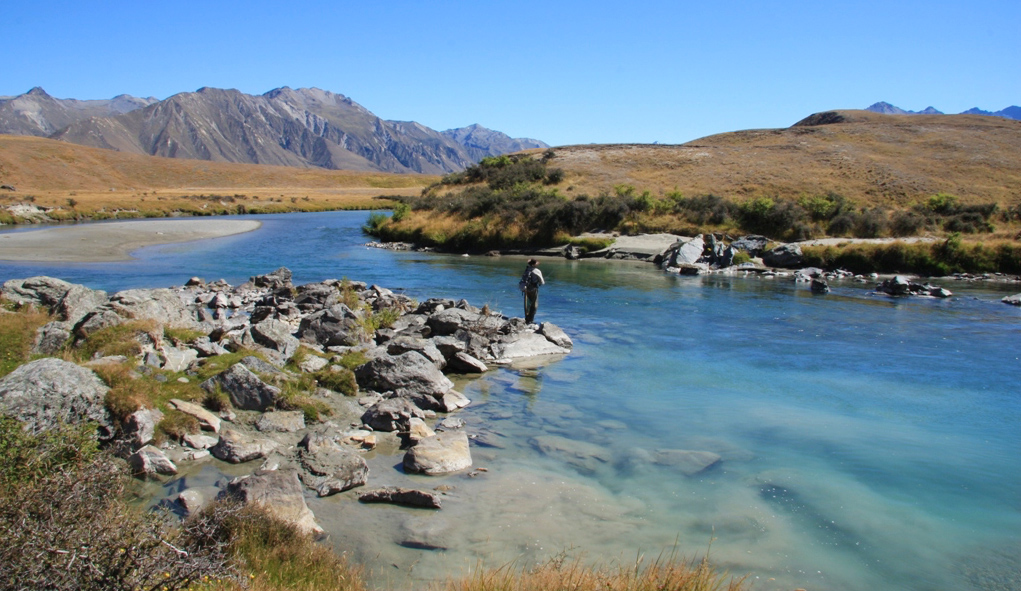 fly fisherman stands looking for trout in wide view of Ahuriri River with valley in background