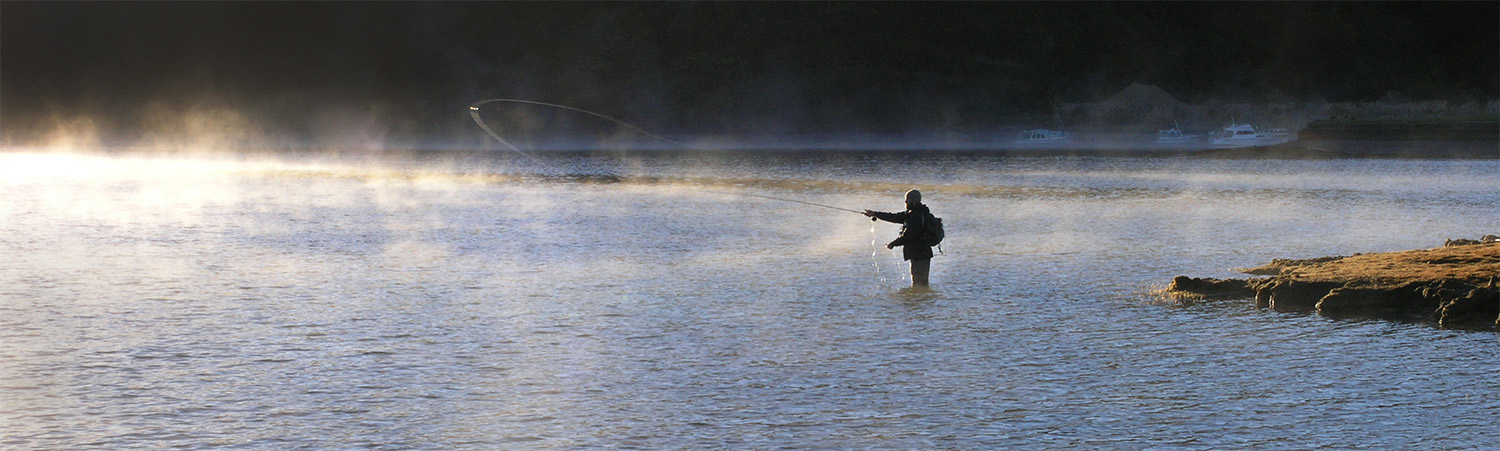 man fly fishing on a lake at sunrise, light illuminating mist on the water, New Zealand