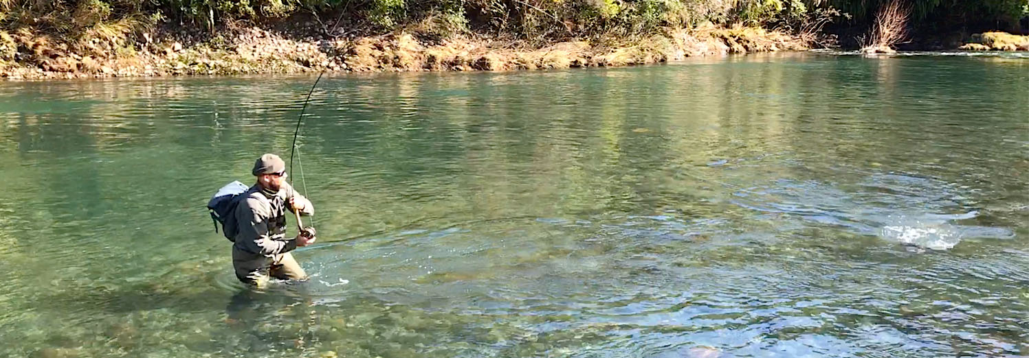 Fly fisherman in a Tongariro river pool, wearing Traper Book Waders holding a trout rod bent as there's a trout on splashing in the water